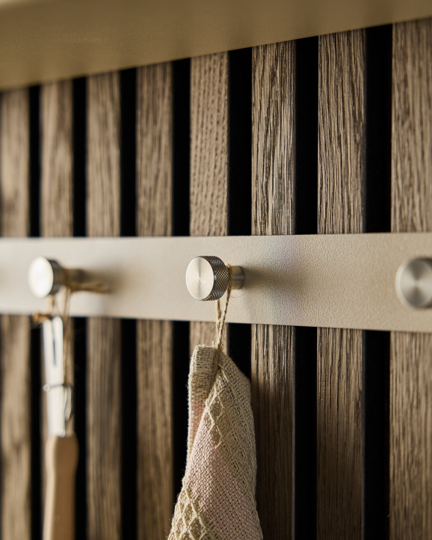 Close-up van de WoodUpp Norr kapstok (Beige/roestvrij staal Steel, X5), gemonteerd op een houten lattenwand Akoestisch paneel, met een handdoek en een gereedschap met handvat eraan.