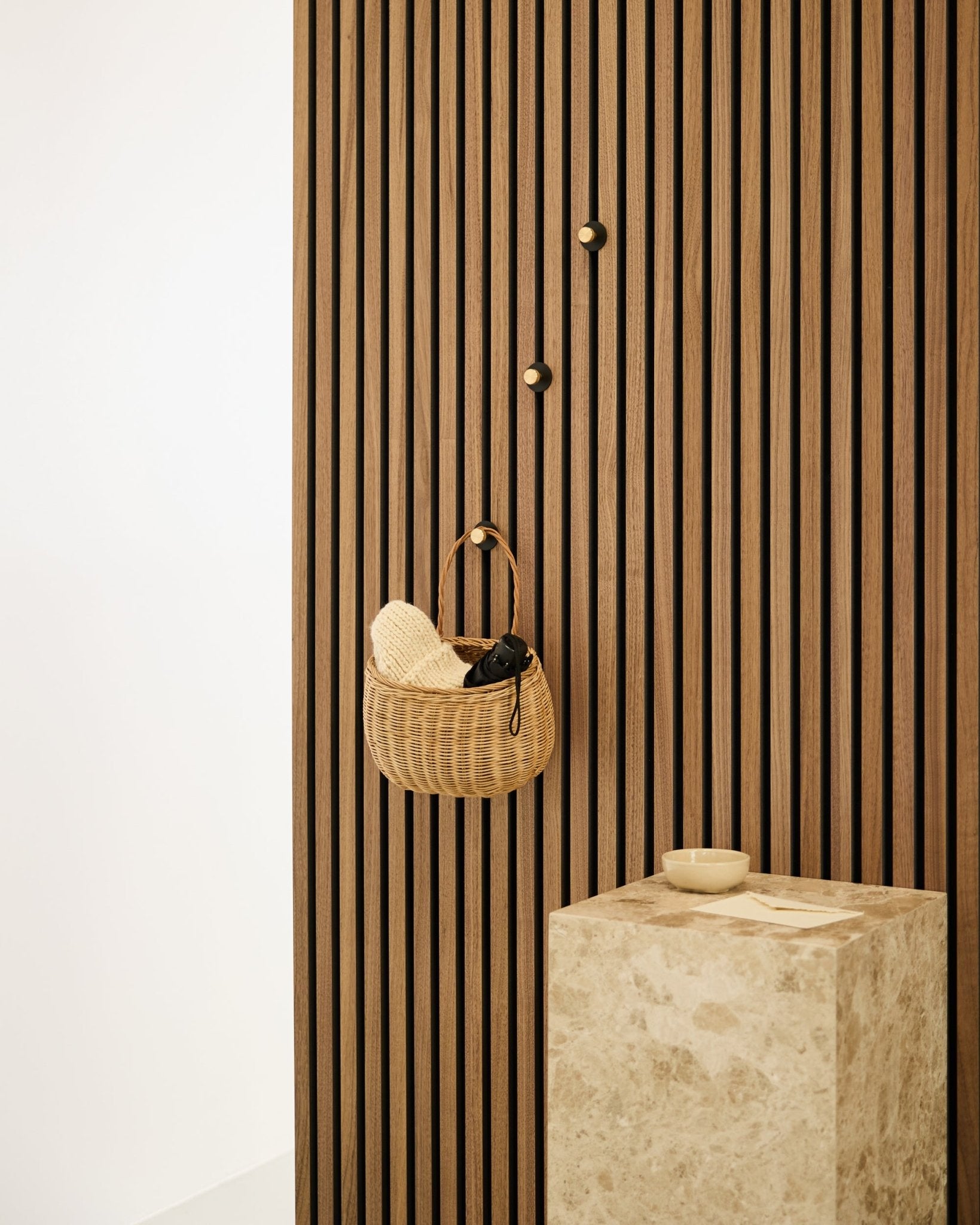 Norr kapstokhaak van WoodUpp gemonteerd op een Scandinavische Akupanel akoestische lattenwand, naast een beige stenen sokkel met een small schaal in een minimalistisch interieur.
