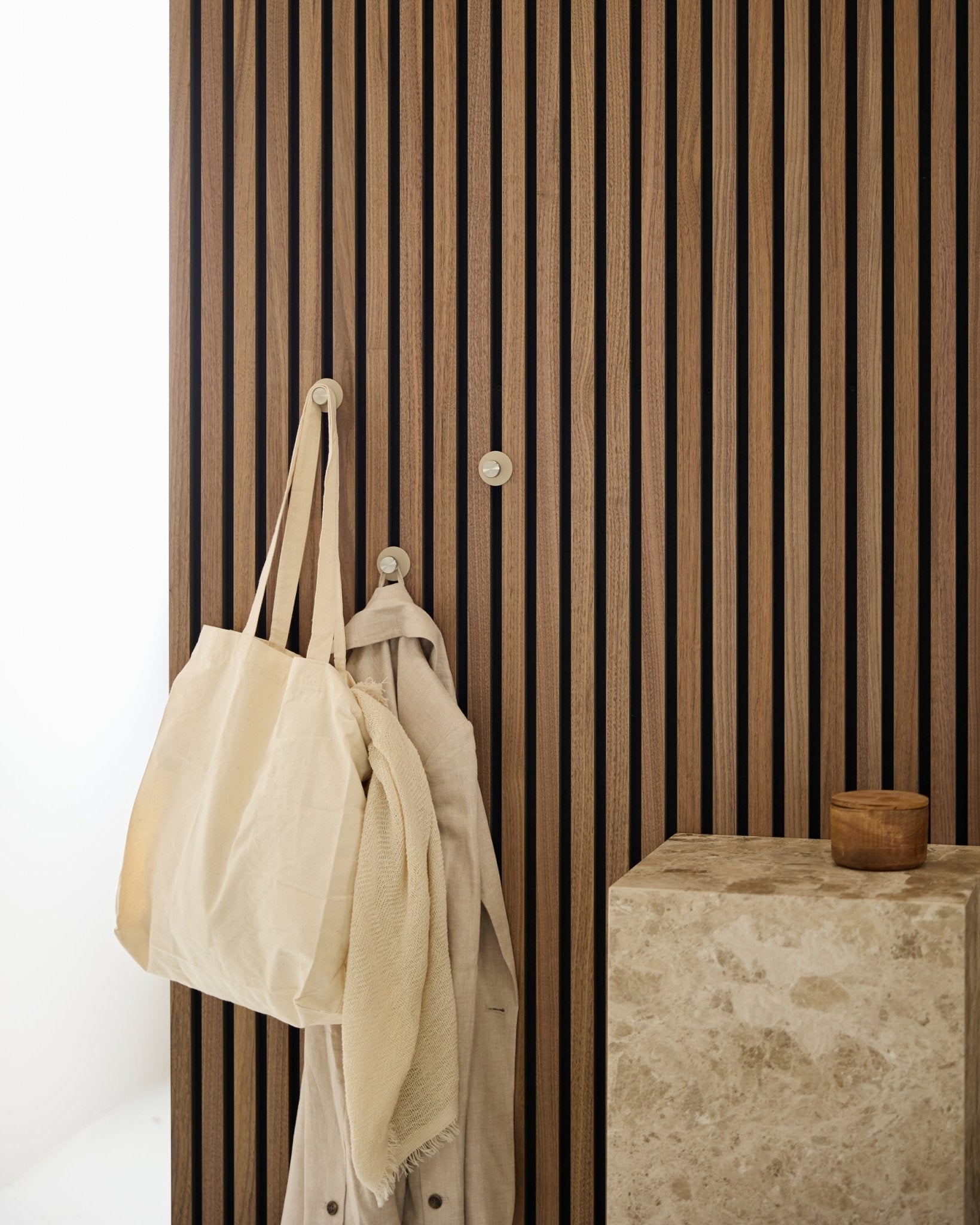 Een beige draagtas en een lichte stof hangen aan een WoodUpp Norr kapstokhaak (Beige/roestvrij Steel), gemonteerd op een vertical Akupanel Akoestisch paneel naast een stenen oppervlak.