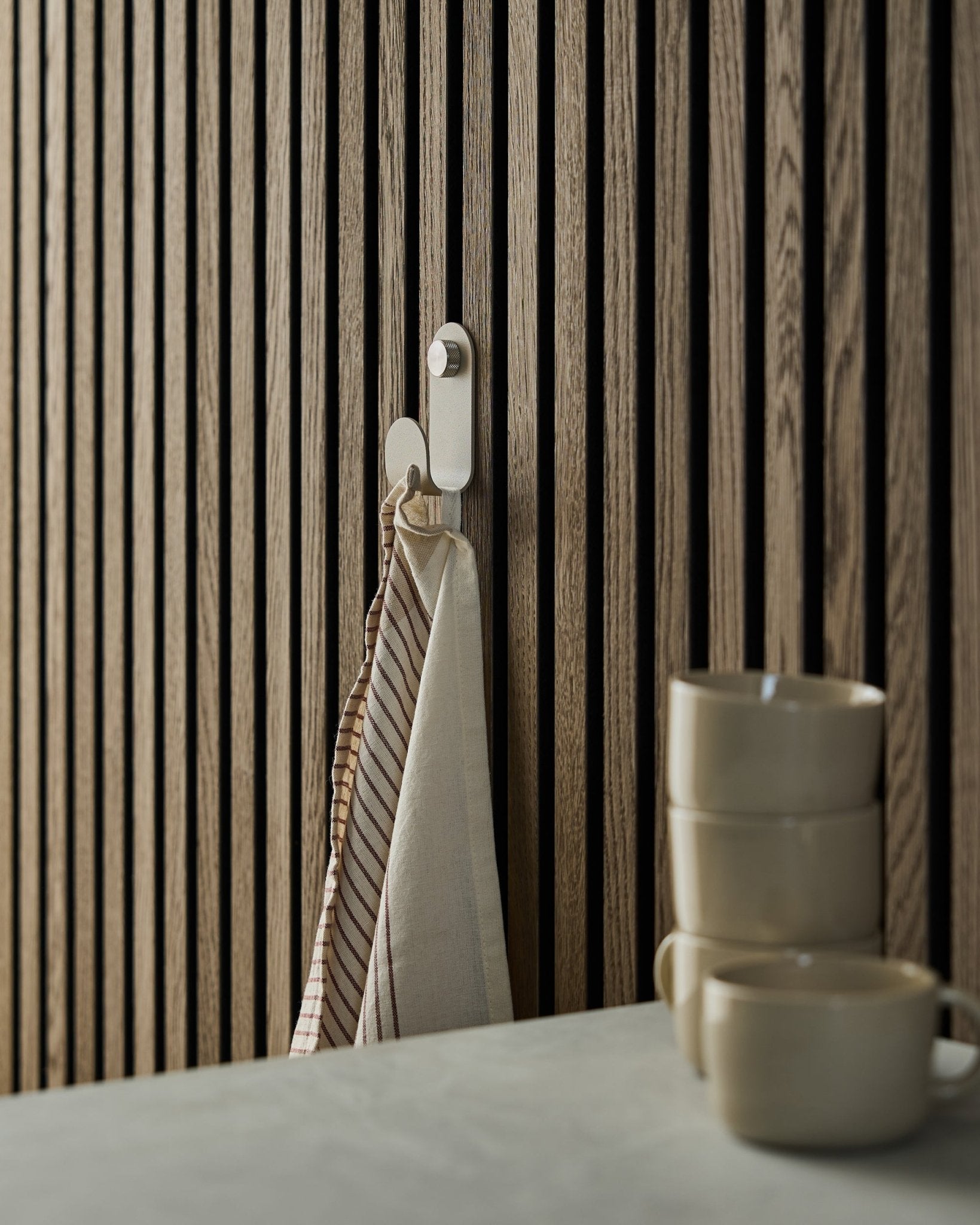 Een gestreepte handdoek hangt aan een WoodUpp Norr Single Hanger in beige, gemonteerd op een akoestisch lattenwandpaneel in Scandinavische stijl; ervoor staan drie beige mokken opgestapeld.