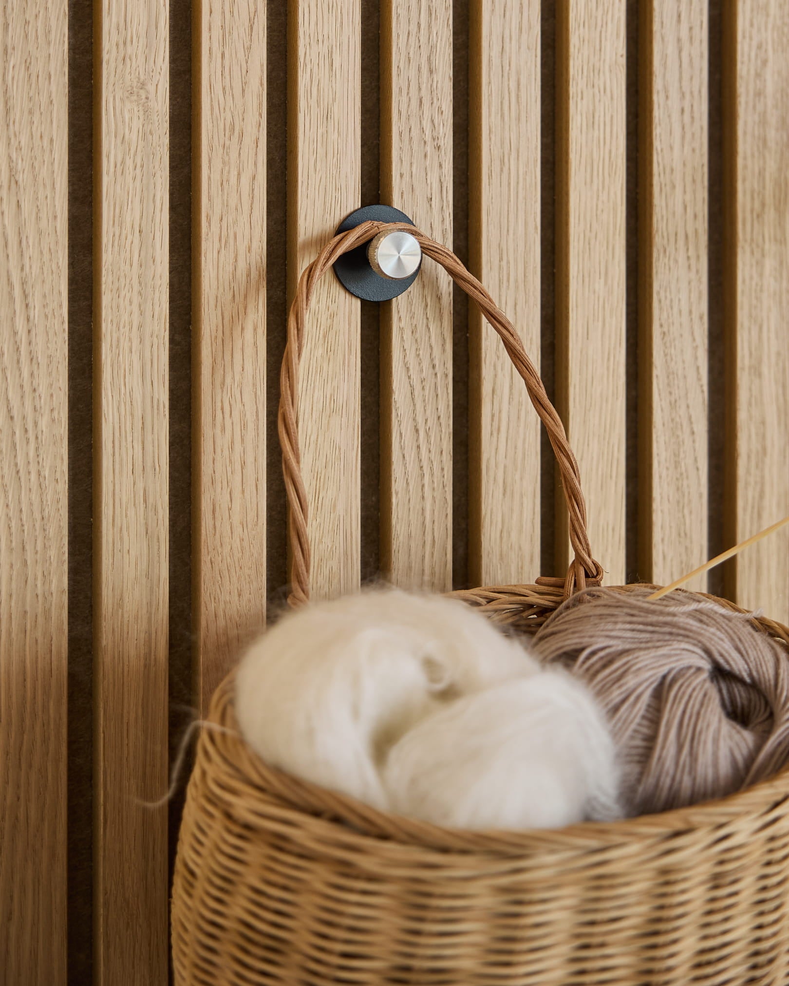 Een witte en beige gevlochten mand van garen hangt aan een Norr Coat Hook / X1, Black/Stainless Steel van WoodUpp, gemonteerd op een Akupanel akoestisch lattenwandpaneel.