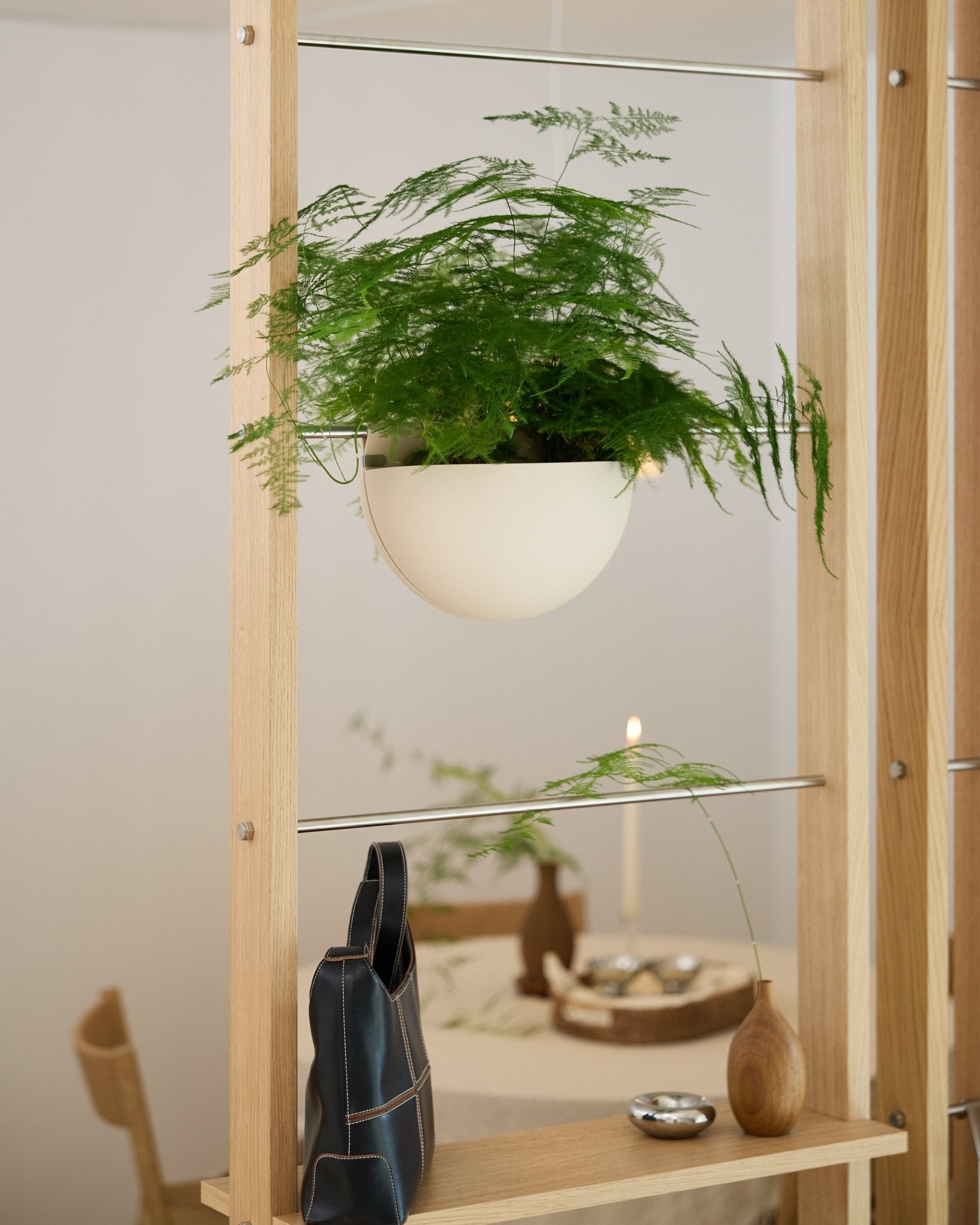 Een groene varen in een lichtbeige WoodUpp Gro Soft Pocket BM2-plantenbak staat op een houten plank boven een zwarte tas, een vaas en een gedekte eettafel.