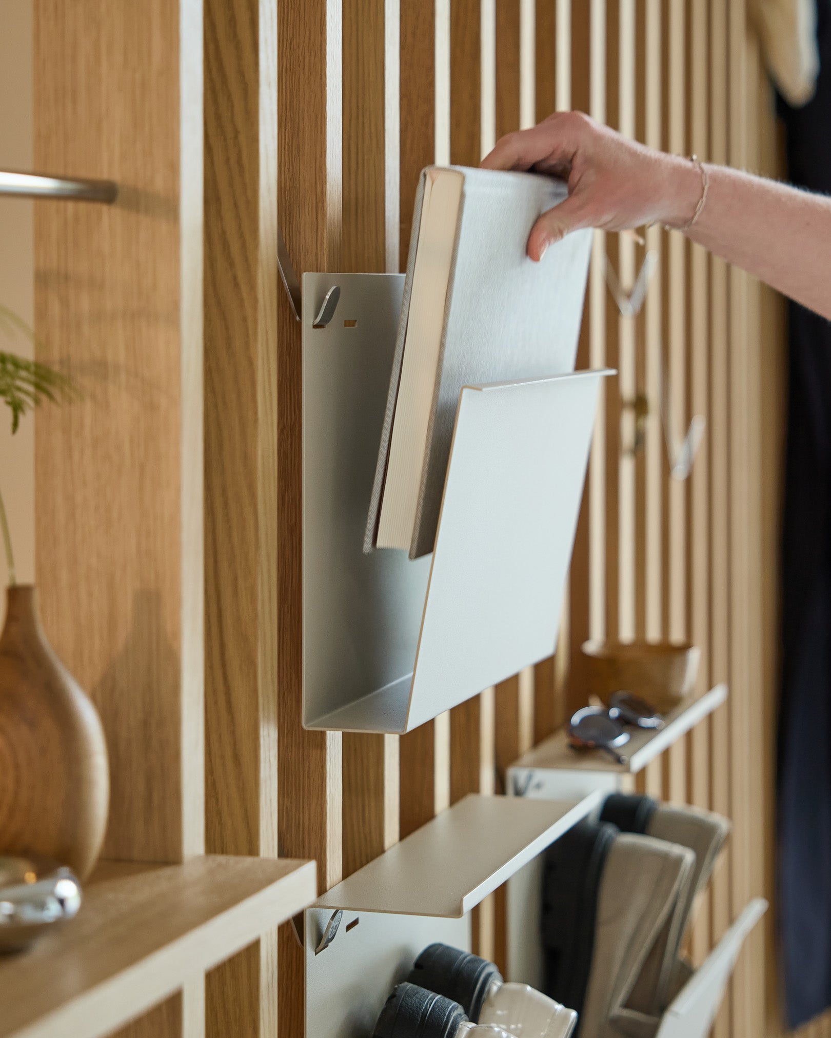 Een hand zet Nell | Magazine Storage / Beige van WoodUpp op een gepoedercoate steel plank, gemonteerd op een Nordic houten Akoestisch paneel, met items ernaast.