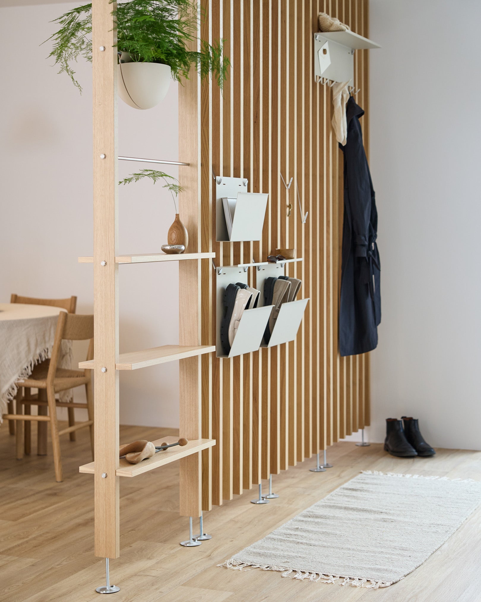 WoodUpp Nell Magazine Storage in beige geïntegreerd in een room divider met steel planken, opbergbakken en kapstokken; eetruimte en vloerkleed zichtbaar op de achtergrond.