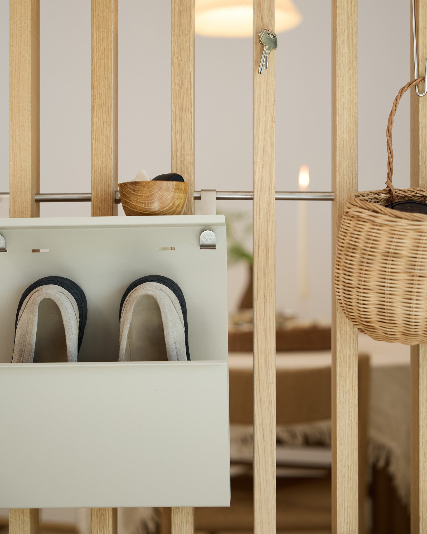 WoodUpp Nell Beige Schoenenopberger toont pantoffels, een kom en een rieten mand op een houten roomdivider; eetruimte zichtbaar op de achtergrond.