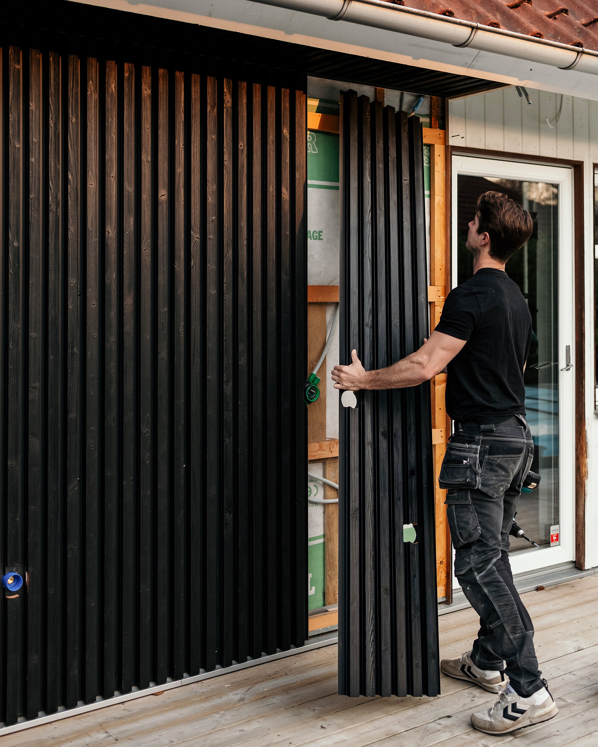 Een man monteert verticale zwarte Aluwood-gevelpanelen op de buitenmuur van een huis terwijl hij op een pas gebouwd terras staat.