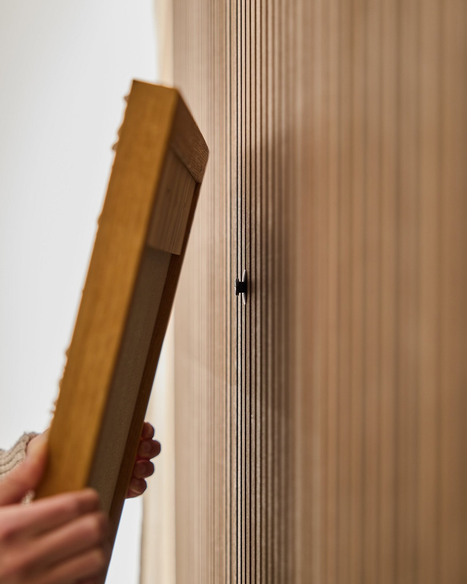 Iemand gebruikt de WoodUpp Dupps | Original set om een frame voor een Akupanel akoestisch houten paneel te monteren, met focus op het bevestigen van een small black haak bij vertical latten.
