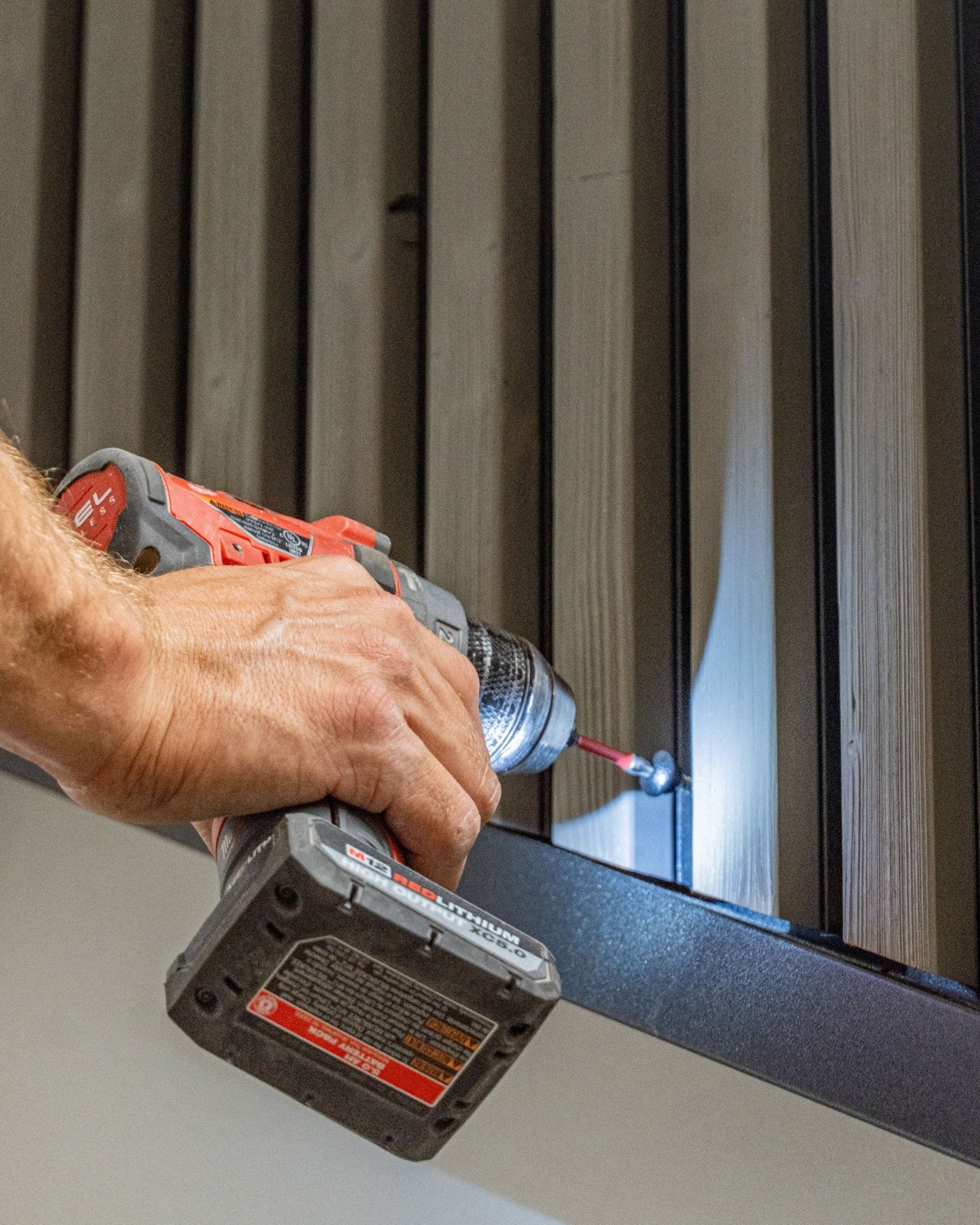 Een persoon bevestigt een black WoodUpp Front Screw (4.8x35, Ø9.5-ring) in een vertical Aluwood-gevelpaneel met een accuboormachine.