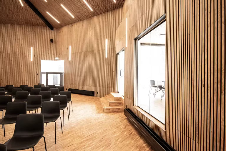 Ruime vergaderruimte met akoestische houten panelen van Akupanel aan de muren en het plafond, rijen zwarte stoelen, grote ramen en verticale verlichting.