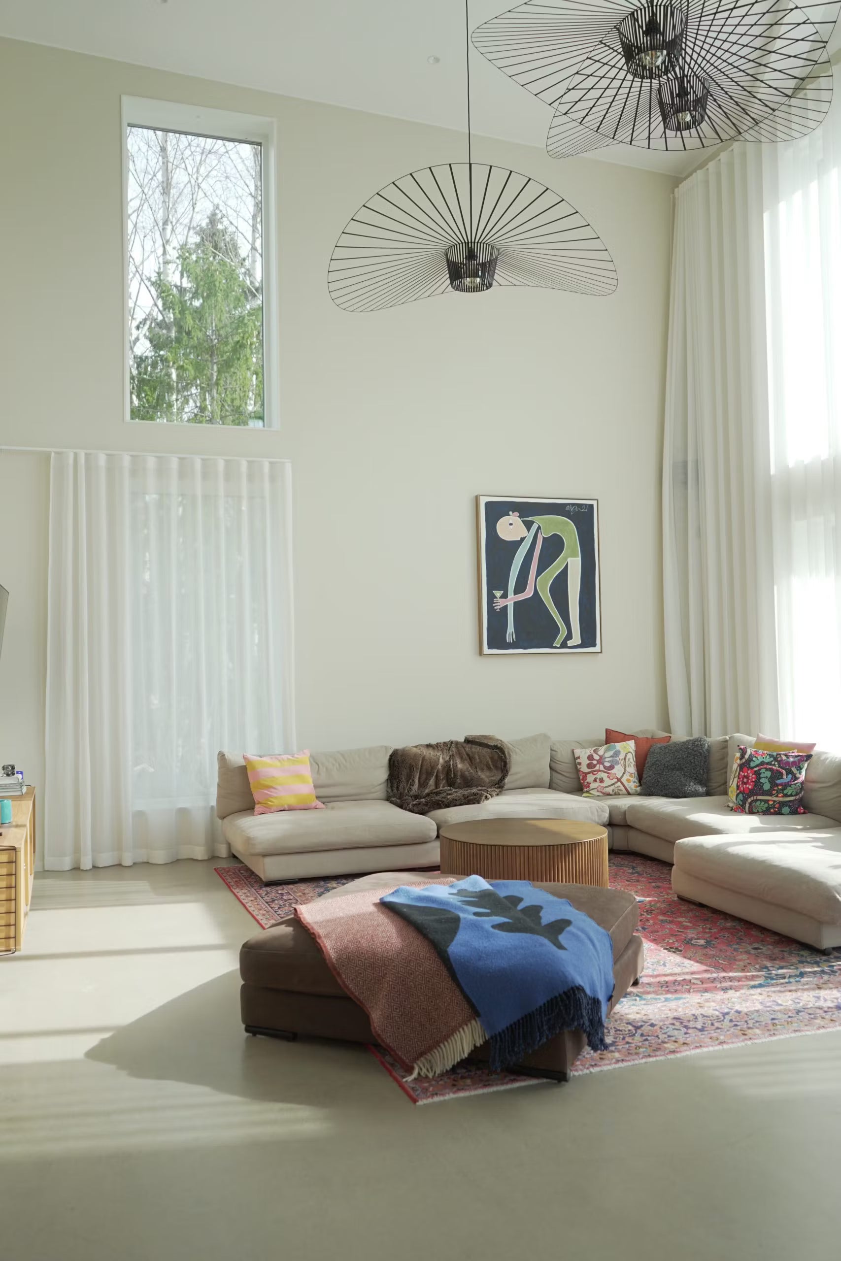 Black wire-frame pendant lights over a beige living room with tall windows and sheer white curtains.
