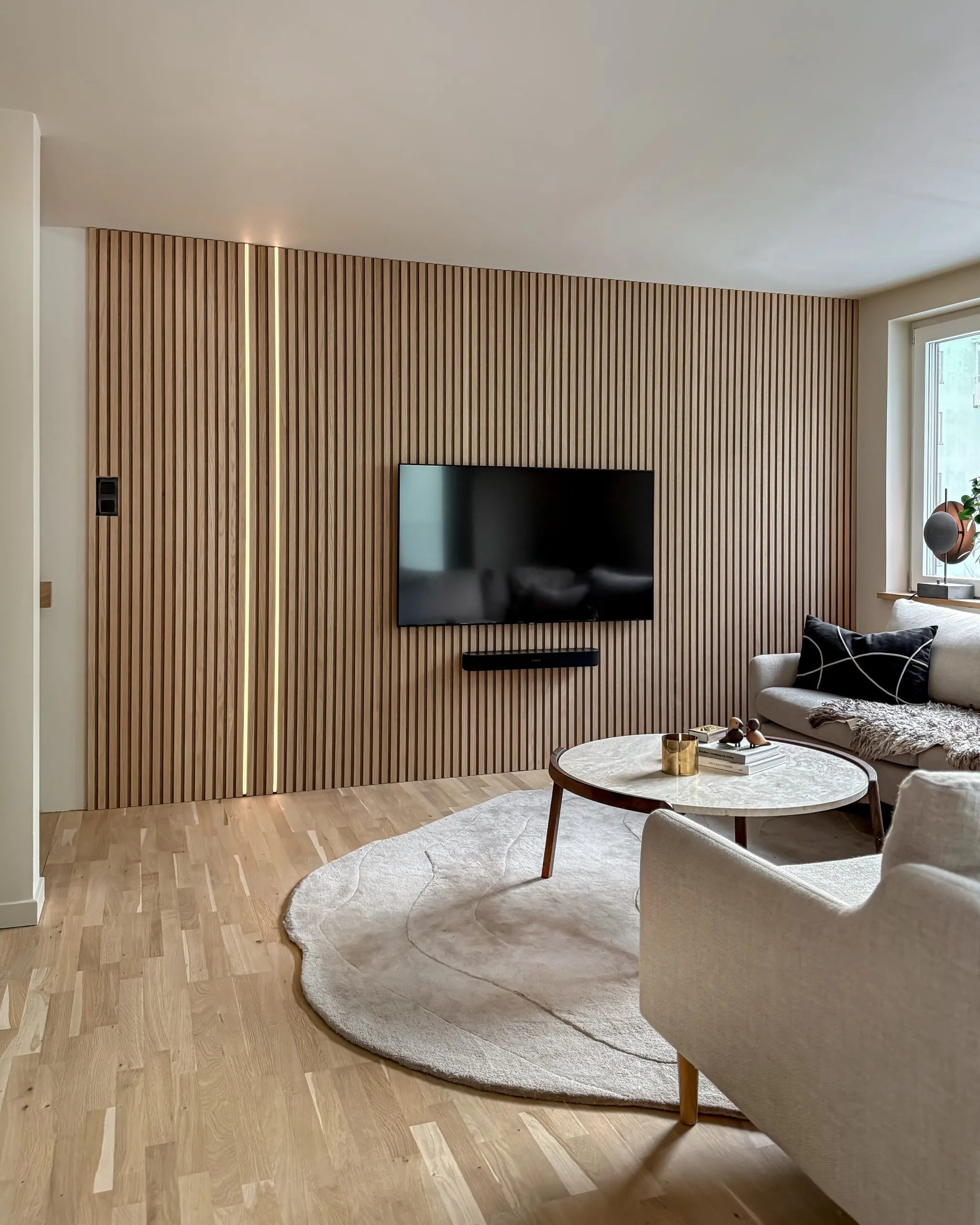 Moderne woonkamer met een Scandinavische Akupanel akoestische houten latten accentmuur, gemonteerde tv, ronde salontafel, crèmekleurige bank, groot vloerkleed op lichte houten vloeren.