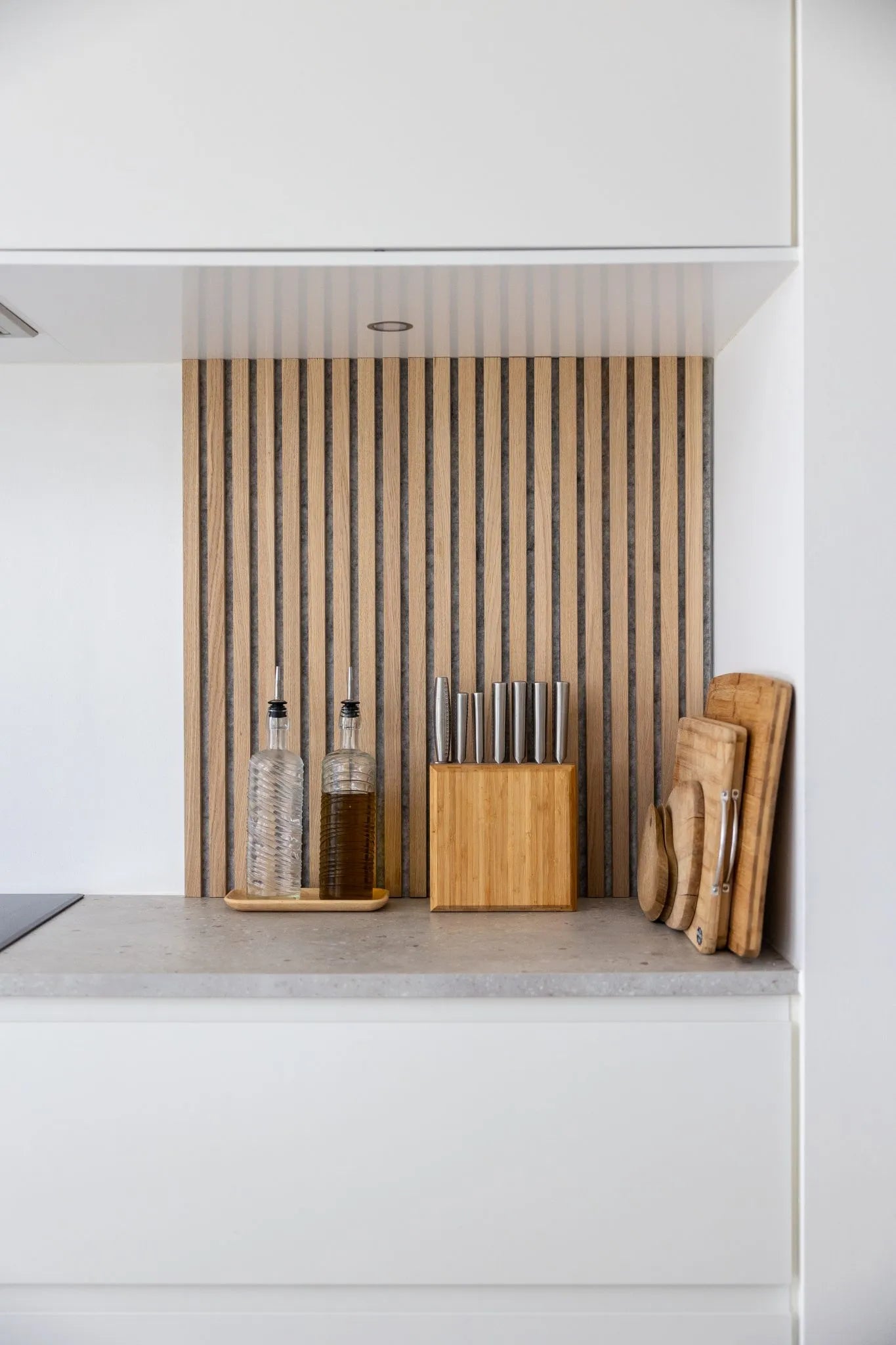 Modern aanrecht met olieflessen, houten messenblok en gestapelde snijplanken tegen een Scandinavische Akupanel akoestische houten lamellenachterwand en witte kasten.