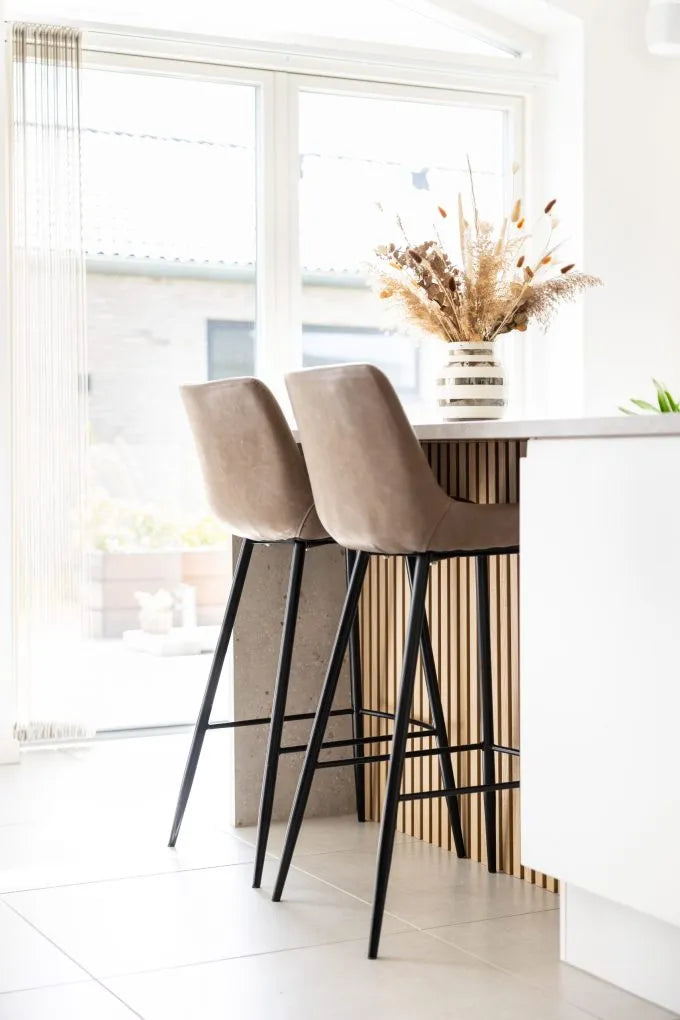 Beige barkrukken bij een keukeneiland met een vaas gedroogde bloemen, grote ramen en een Scandinavische eigentijdse stijl met akoestische panelen op de muren.