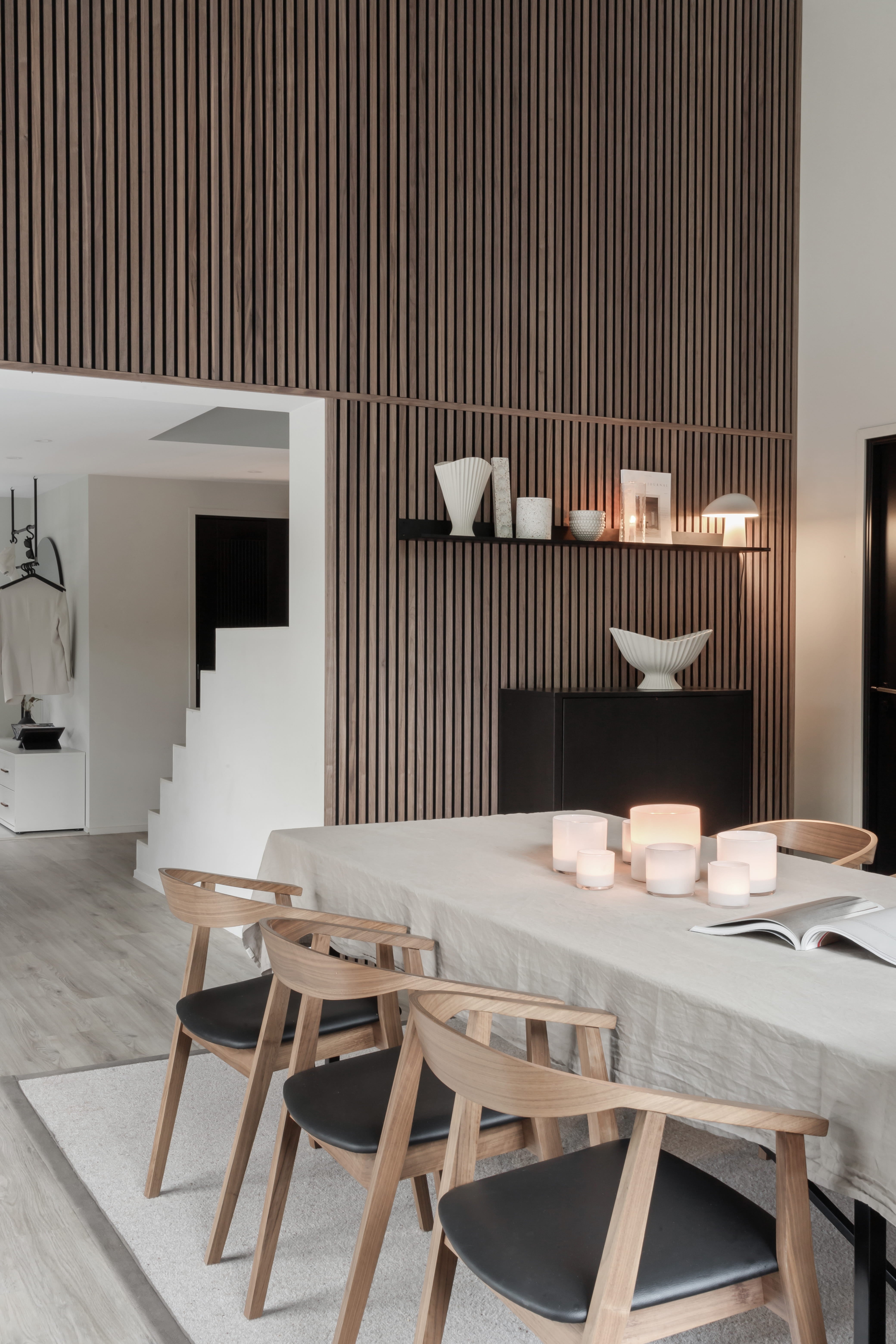 Moderne eetkamer met een lichthouten tafel, vier stoelen, kaarsen en een minimalistische inrichting. Verticale Akupanel akoestische houten lattenwand en open trap in zicht.