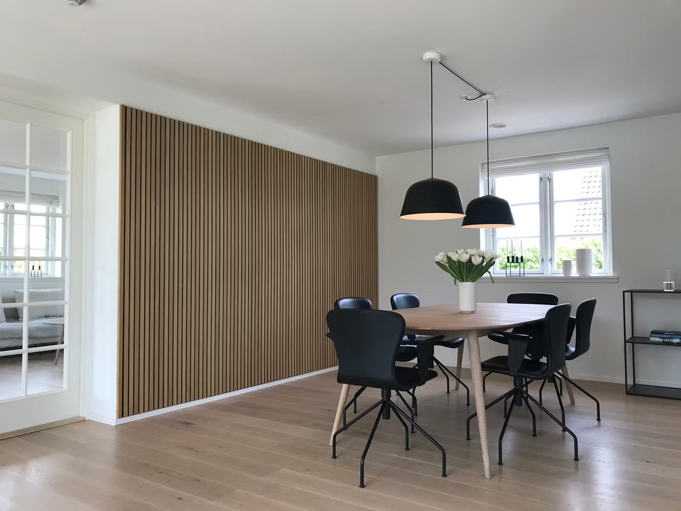 Moderne eetkamer met houten vloer, akoestische Akupanel lamellen, zwarte stoelen, houten tafel, hanglampen en een wit bloemstuk in het midden.