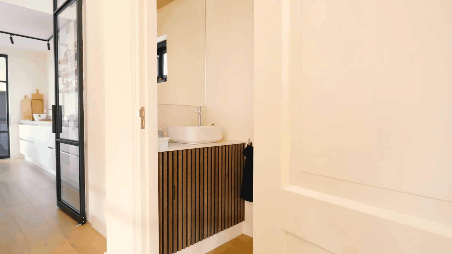 Moderne badkamer met ronde wastafel, akoestisch badmeubel met houten lamellen, grote spiegel en witte deur naast een lichte hal en moderne keuken.
