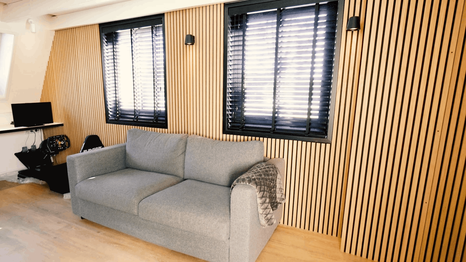 Grijze sofa met gedessineerde deken geplaatst voor een Scandinavische Akupanel akoestische houten lattenwand, geflankeerd door twee ramen met zwarte jaloezieën en een compact bureau.