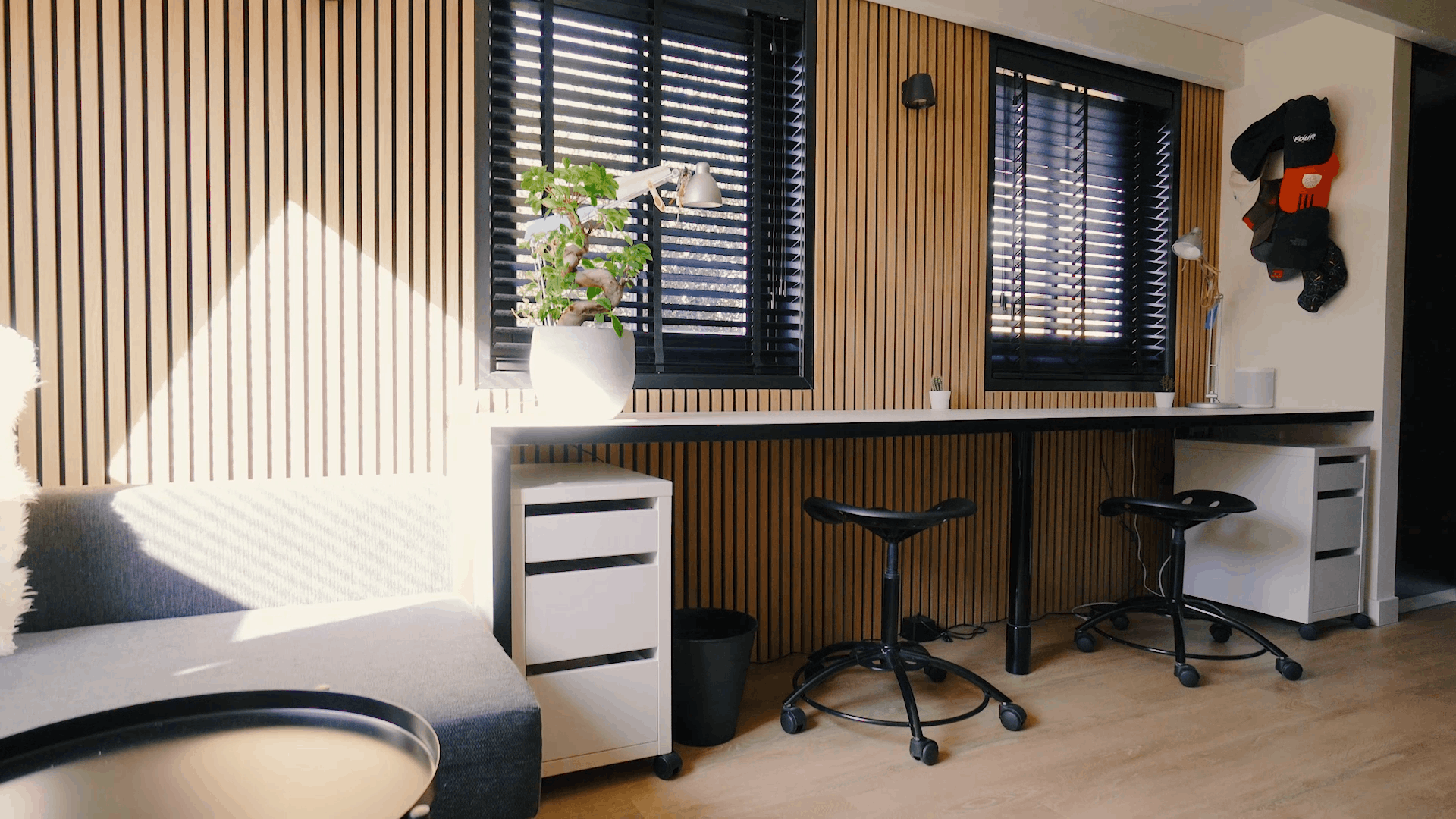 Modern kantoor met Scandinavische akoestische houten panelen op de muren, ergonomische werkplekken, zwarte krukjes, een plant in potten en zonlicht door zwarte jaloezieën.