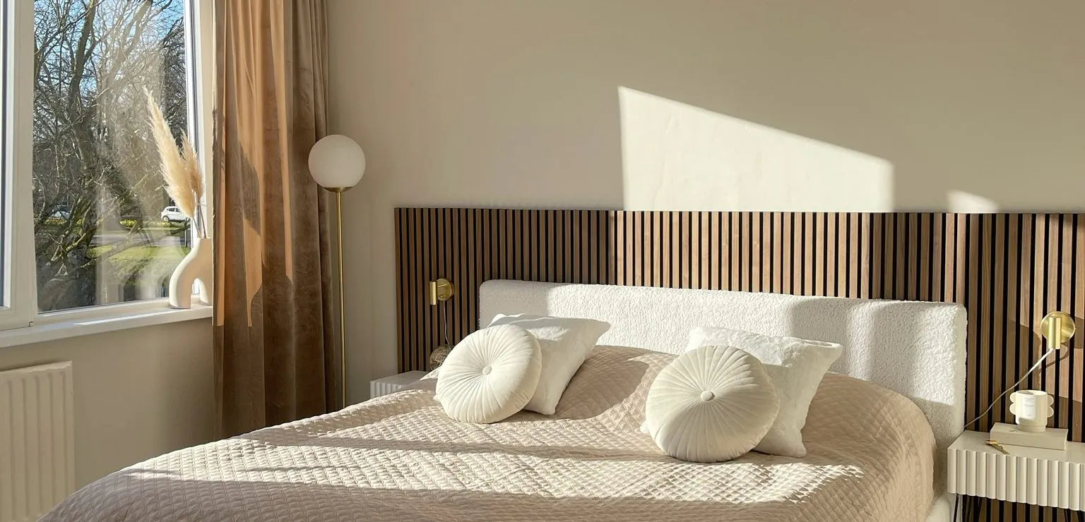 Zonovergoten moderne slaapkamer met een beige gewatteerd bed, ronde kussens, Akupanel houten latten hoofdeinde en minimale inrichting naast een groot raam met transparante gordijnen.