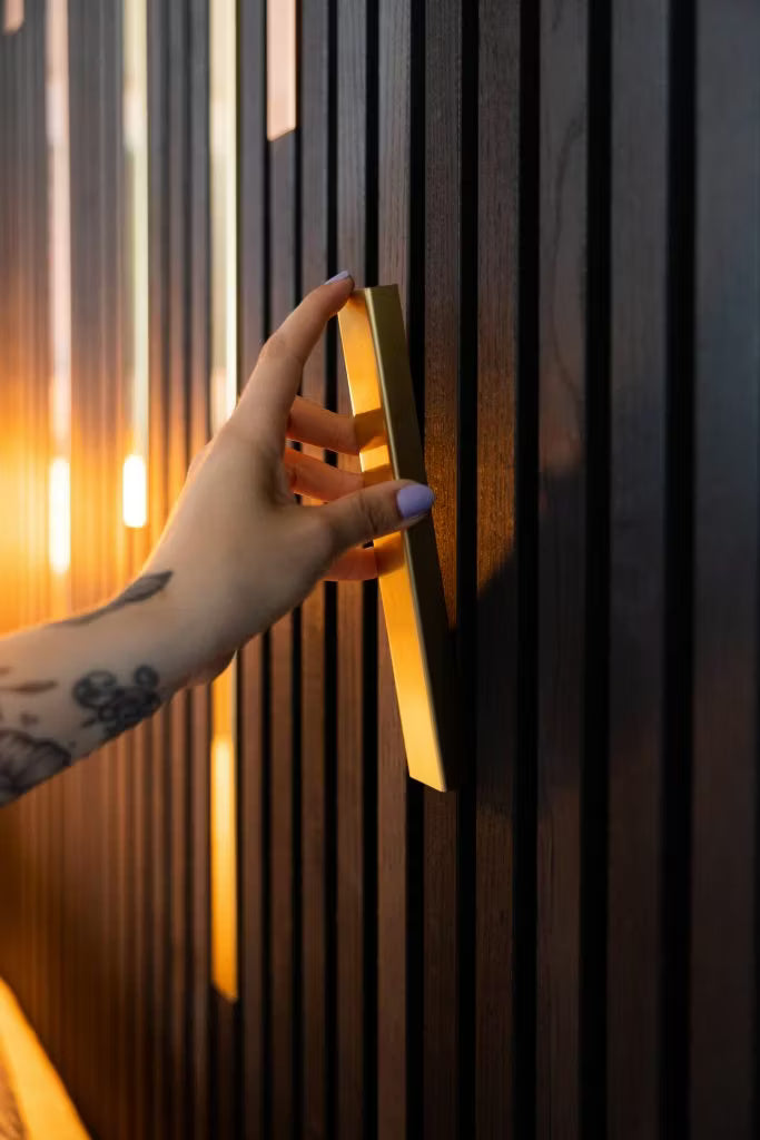 Een getatoeëerde hand met lichtpaarse nagels houdt een gouden handvat vast op een strakke, donkere wand met akoestische houten panelen van Akupanel, benadrukt door warme verlichting.