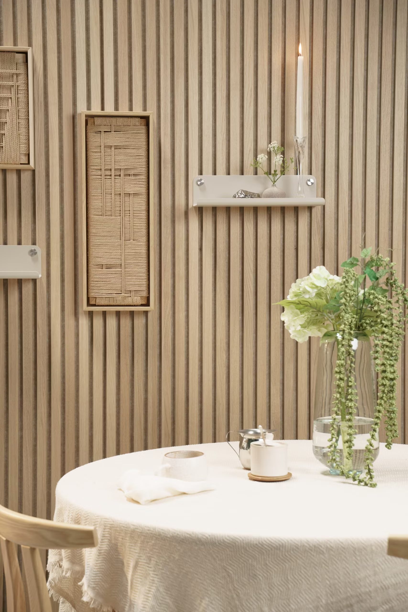 Een ronde tafel met een licht tafelkleed, verse bloemen in een vaas en een kaars staat voor een Akupanel akoestische houten wand in Scandinavische stijl met minimale planken.