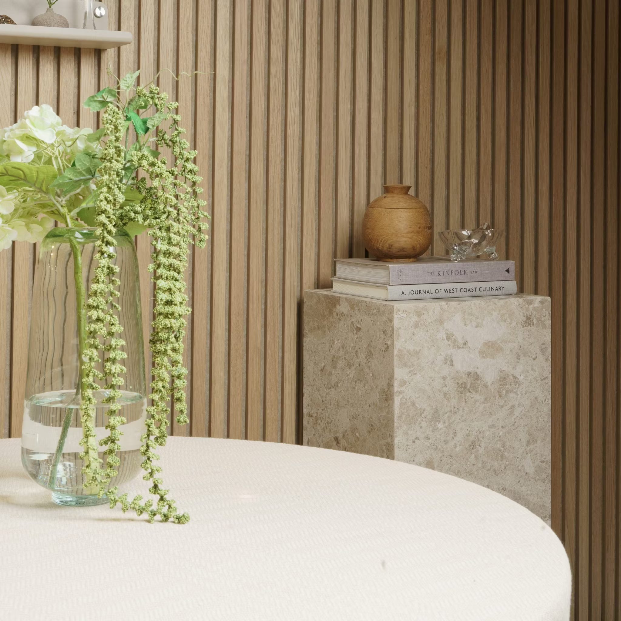 Een vaas met bloemen staat op een ronde tafel; boeken en aardewerk rusten op een marmeren sokkel tegen een Akupanel akoestische houten paneelmuur.