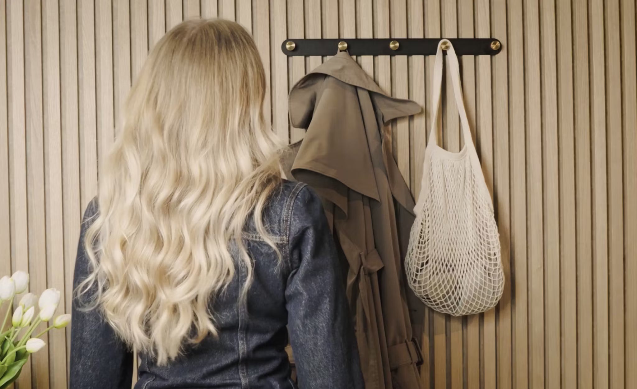 Een vrouw met lang blond haar staat voor een kapstok met een trenchcoat, witte netzak en verse witte tulpen naast een akoestisch houten paneel van Akupanel in de hoek.