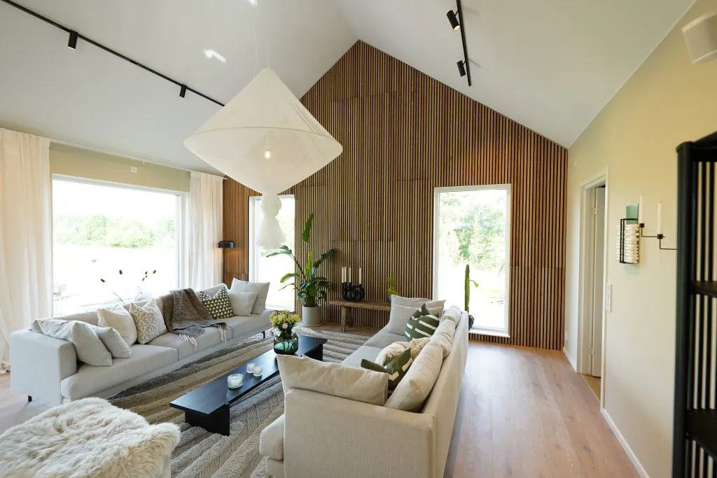 Moderne woonkamer met hoog plafond, grote ramen, lichte banken, Akupanel akoestische houten accentmuur, kamerplanten en overvloedig natuurlijk licht.