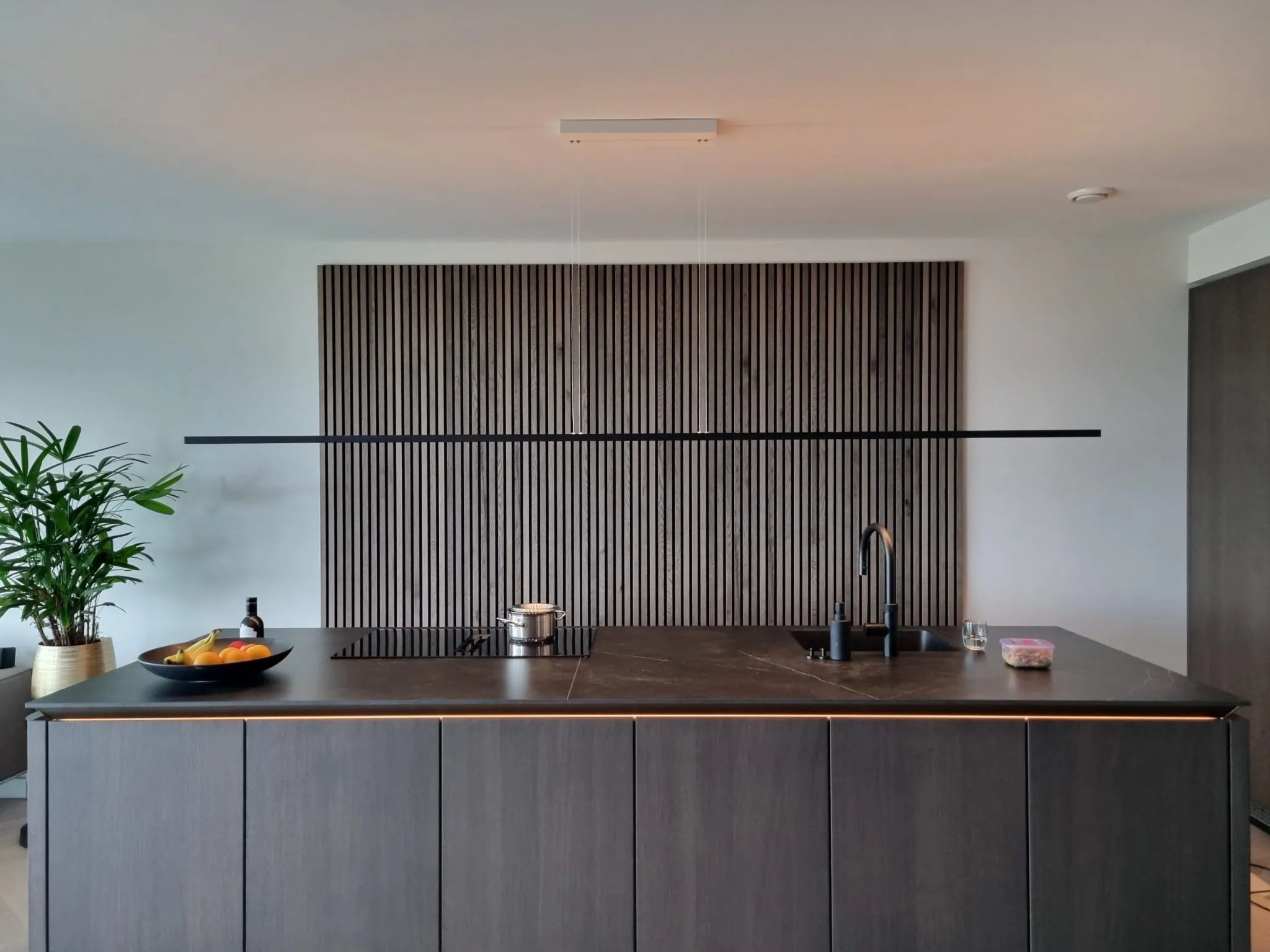Moderne keuken met een donker eiland, ingebouwde gootsteen en fornuis, fruitschaal en plant in pot. Verticale Akupanel akoestische houten panelen accentueren de muur achter het eiland.