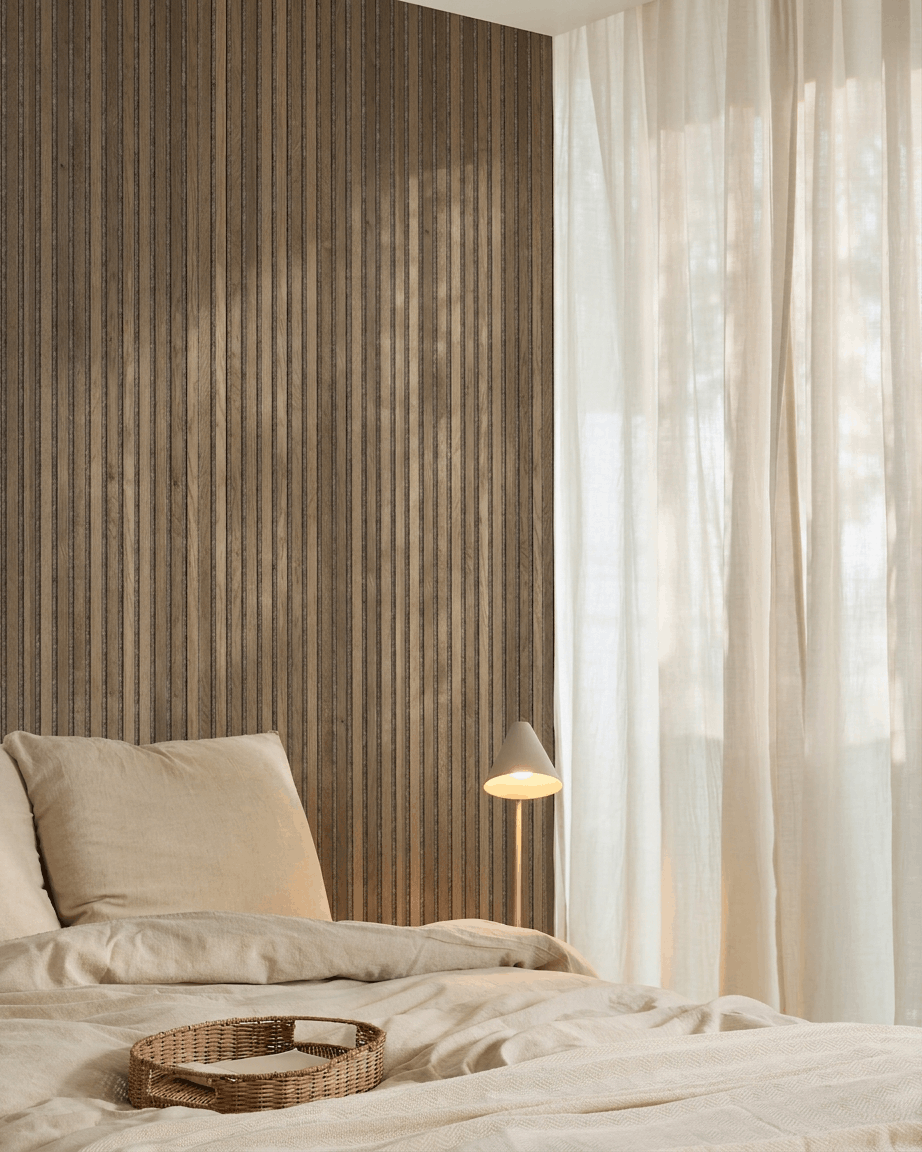 Minimalistische slaapkamer met beige beddengoed, een rieten dienblad, een WoodUpp Akupanel Rustic Grey Oak akoestisch lattenwandpaneel, een witte lamp en vitrage.