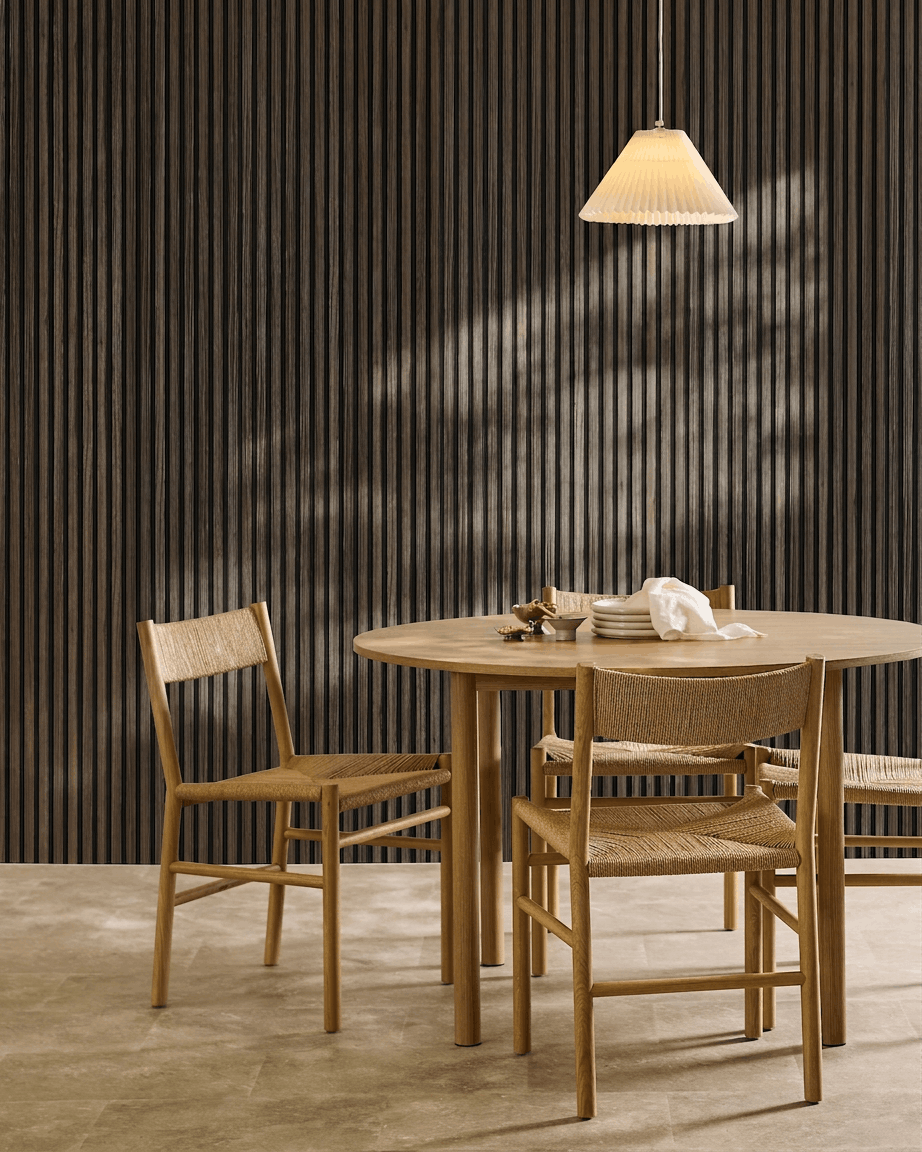 Een ronde tafel met vier gevlochten stoelen staat naast een WoodUpp Akupanel Black Oak Akoestisch paneel, onder een geplooide hanglamp.