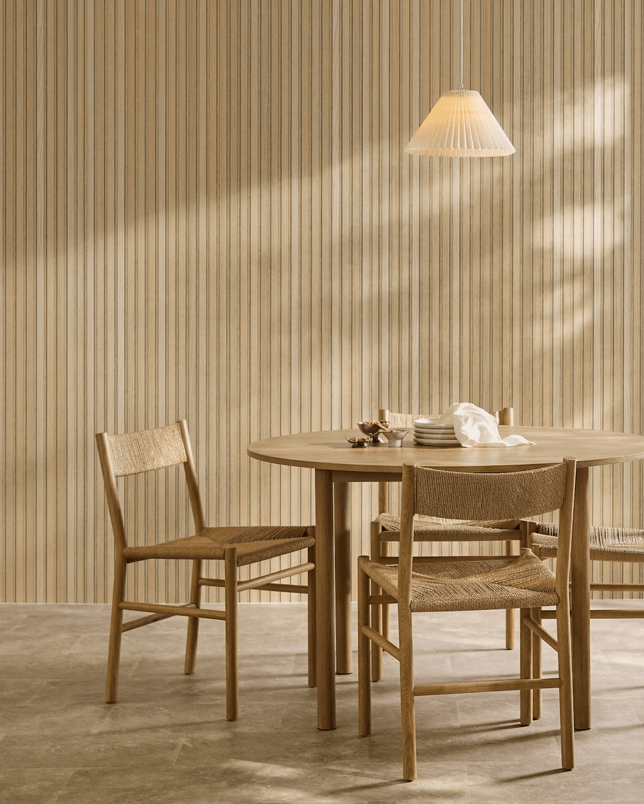 Minimalistische eethoek met een ronde tafel, geweven stoelen en een hanglamp naast een WoodUpp Akupanel akoestisch houten paneel in Classic Oak met beige vilt.