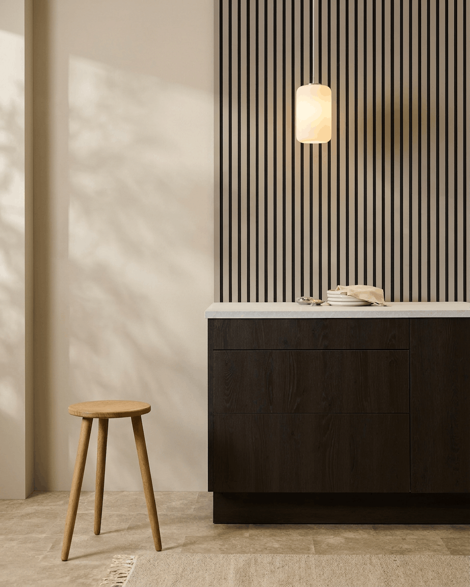 Minimalistische keuken met WoodUpp Akupanel (Stone, Black vilt) akoestische panelen aan de muur, een houten kruk, donkere kasten en een hanglamp in neutrale tinten.