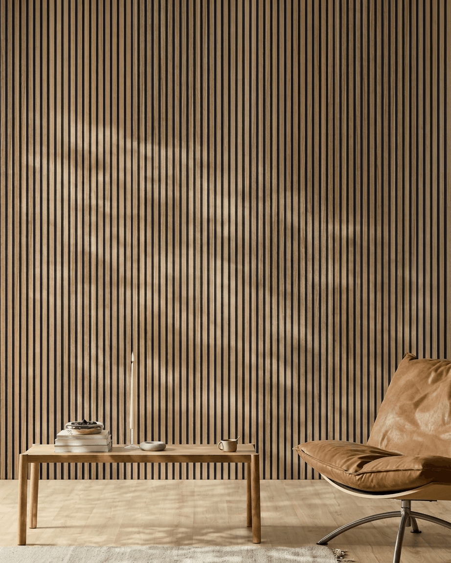 Minimalistische ruimte met een WoodUpp Akupanel Akoestisch paneel (English Walnut-latten, black vilt), een stoel van cognackleurig leer, een tafel van licht hout, een kaars, boeken, een kopje en een wit vloerkleed in het zonlicht.