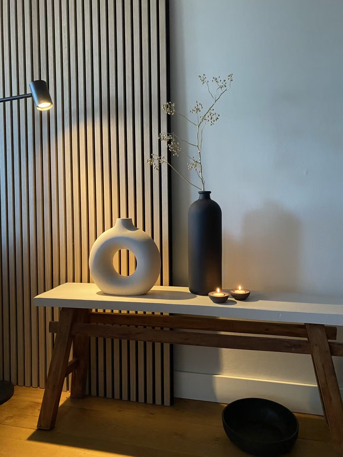 Minimalistisch decor met een witte ronde vaas, een zwarte hoge vaas met gedroogde stengels, twee waxinelichtjes en een zwarte schaal tegen een achtergrond van akoestische panelen van Akupanel-hout.