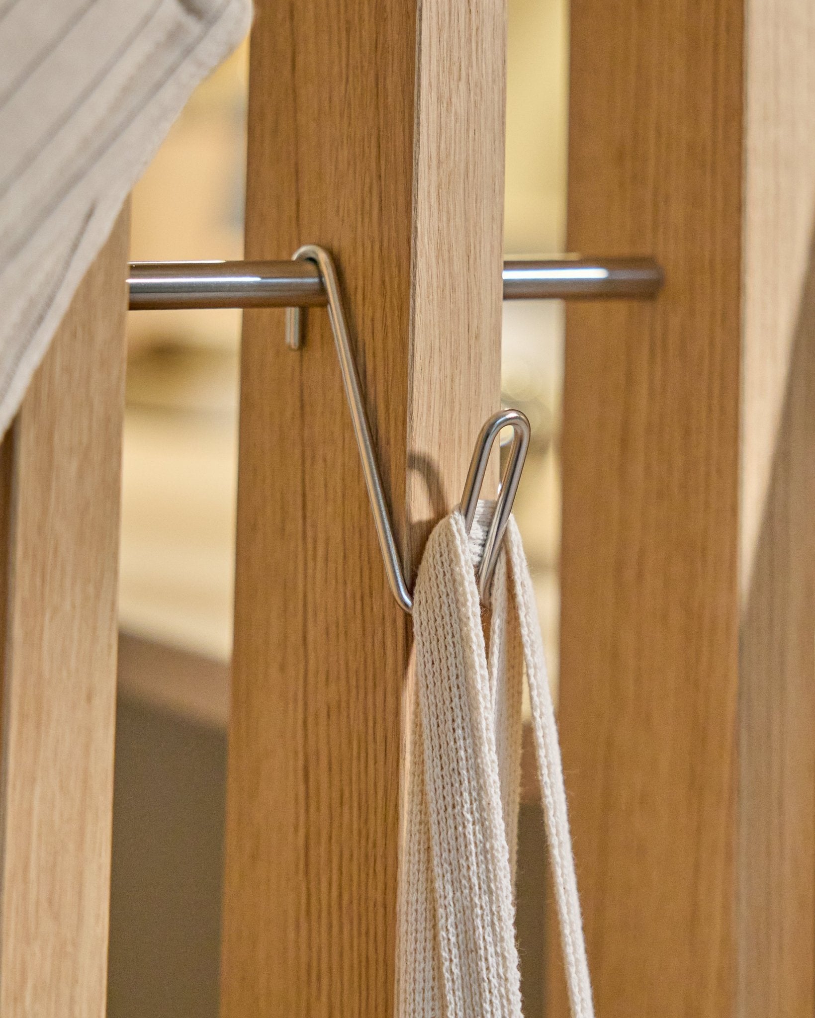 De Alva Wire Hook / BM1 (roestvrij staal Steel, kort) van WoodUpp is gemonteerd op een houten Akoestisch paneel rail en houdt een witte band met metalen stang vast; beige achtergrond.