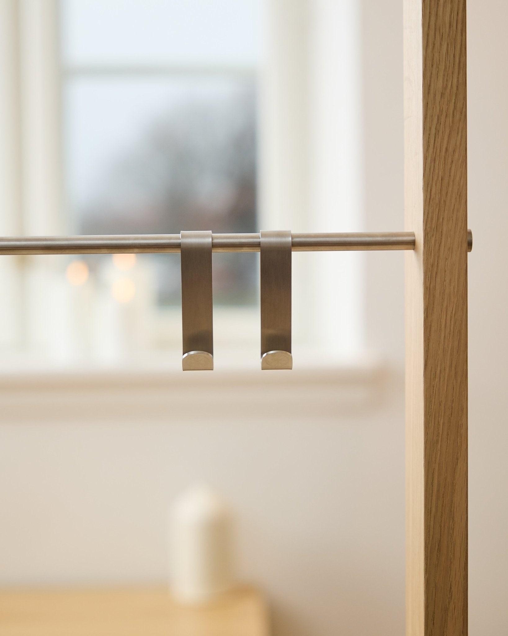 WoodUpp Alva S-Hook BM1  BM2 (2 stuks) in roestvrij steel, hangend aan een stang op een akoestisch houten paneel; op de achtergrond een wazig raam en brandende kaarsen.