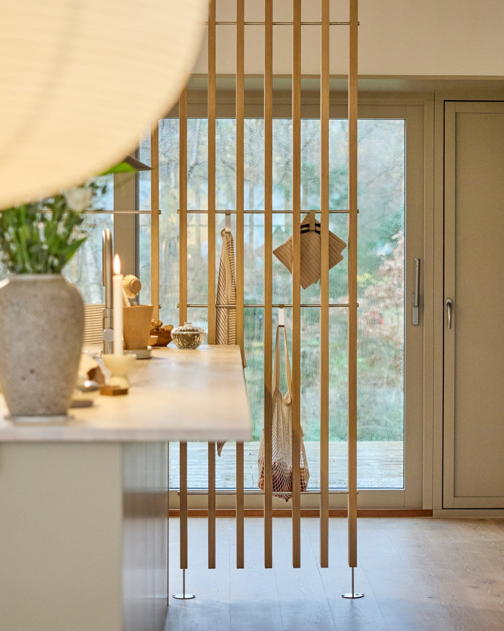 Minimalistische keuken met een WoodUpp Embrace roomdivider (BM1, 286-302 cm) in eikenfineer, met lichte werkbladen, een vaas met bloemen en uitzicht op de tuin.