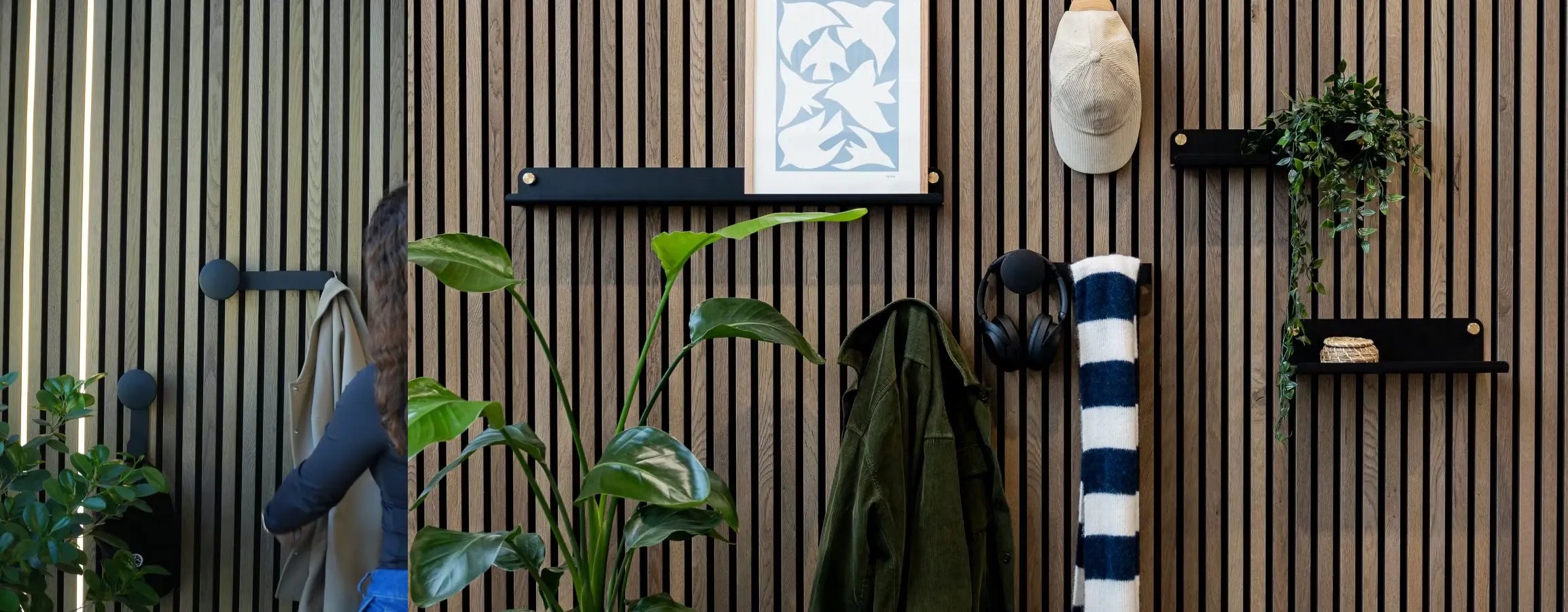 Een man in een blauw jasje loopt langs een Scandinavische Akupanel akoestische houten lattenwand met geïntegreerde planten, planken, kapstokken, koptelefoons en een opgerolde gestreepte handdoek.