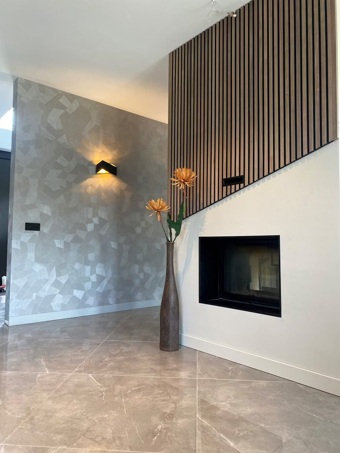 Moderne woonkamer met behang met geometrische patronen, akoestische panelen van Akupanel-hout, een open haard aan de muur en een hoge vaas met oranje bloemen.