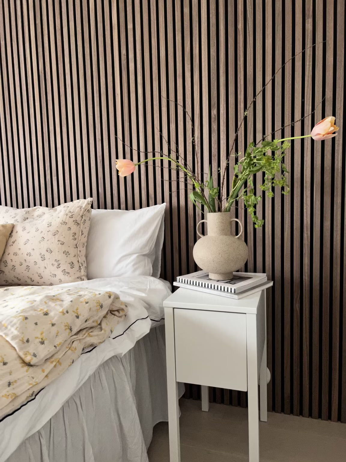 Een slaapkamer met een wit bed met gebloemd beddengoed en een vaas met bloemen, met een Scandinavische houten Akupanel akoestische lamellen accentmuur achter het bed.