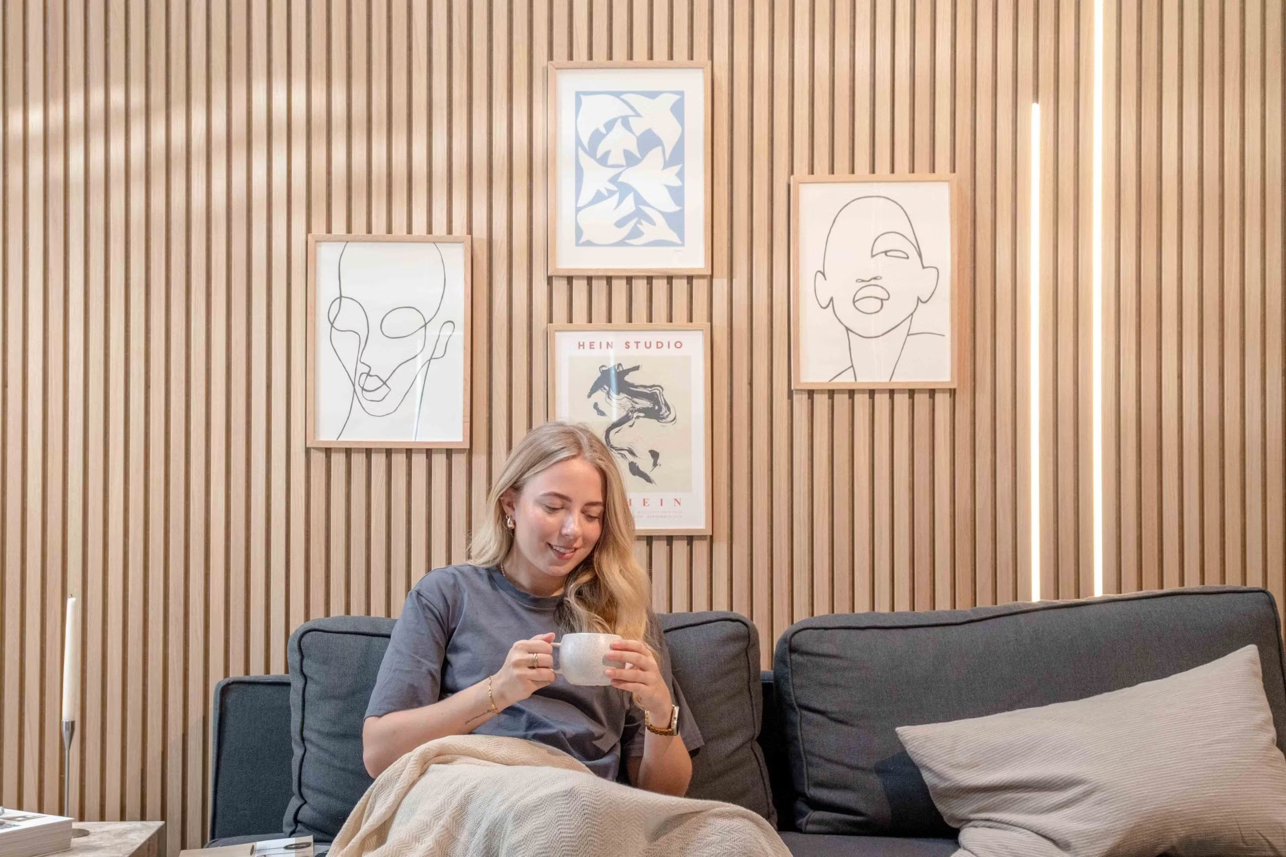 Vrouw ontspant op een bank met een mok, glimlachend onder minimalistische lijnkunst en abstract decor gemonteerd op Scandinavische akoestische slatwallpanelen met textuur.