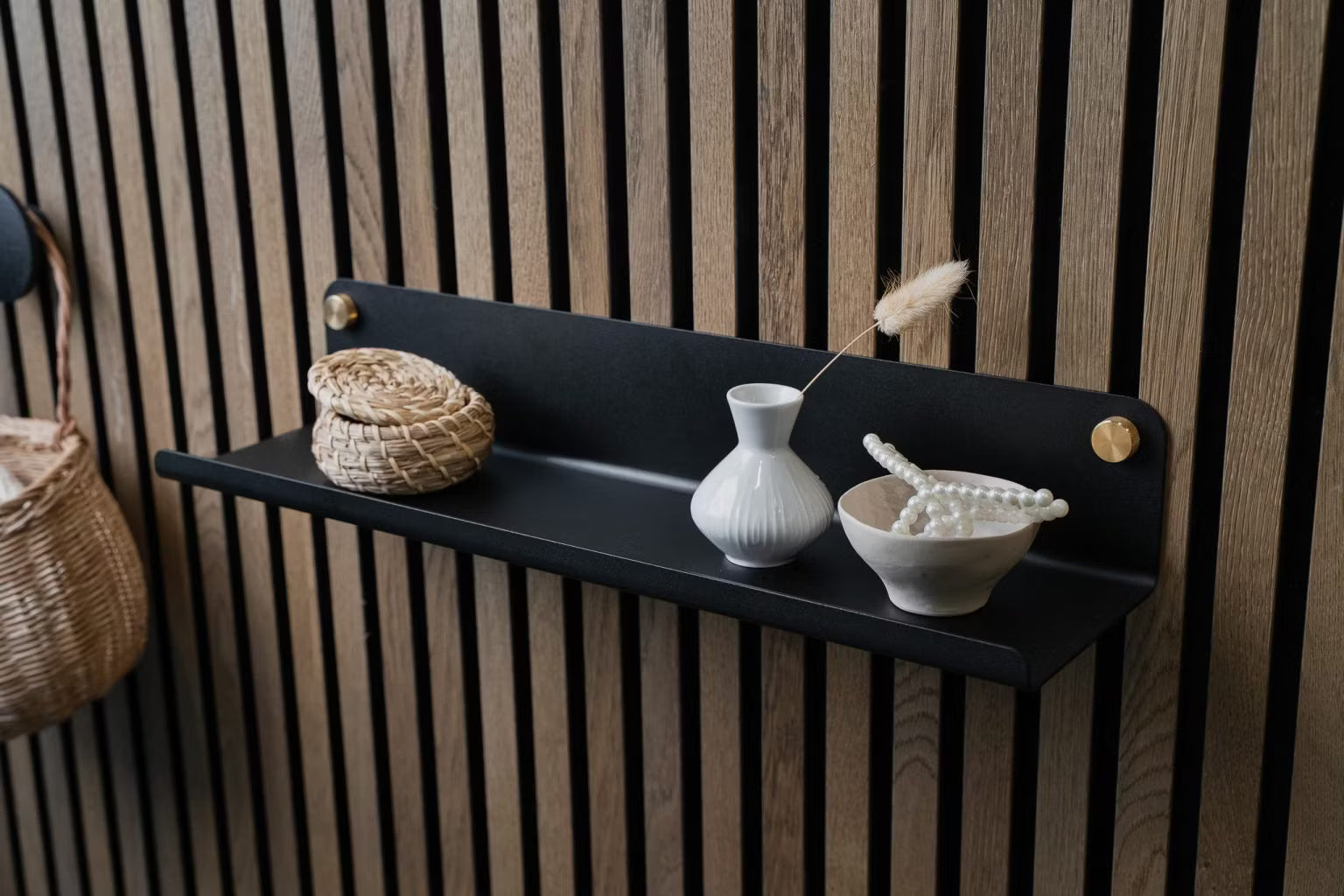 Een zwarte wandplank met een kleine rieten mand, witte vaas en keramische schaal met parels gemonteerd op een Scandinavisch houten akoestisch paneel.